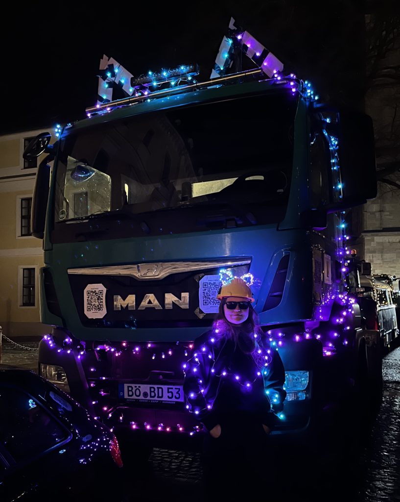 Frau in Pullover mit Lichterkette steht vor LKW mit vielen bunten Lichterketten und Ohren